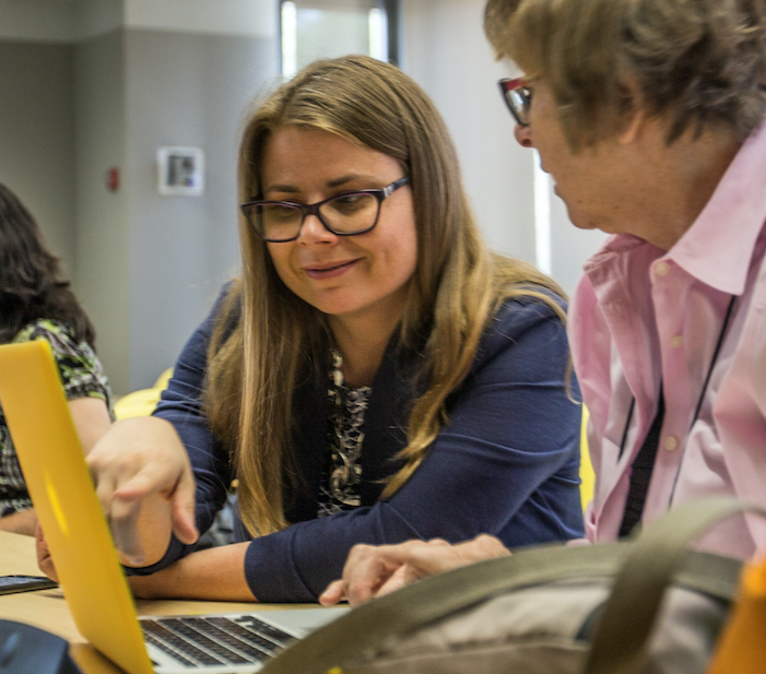 Photo of Laura Gogia pointing at a computer screen while seated next to someone whom she is coaching. This is actually a Wikipedia editing event and not the course coaching referenced in this blog post.