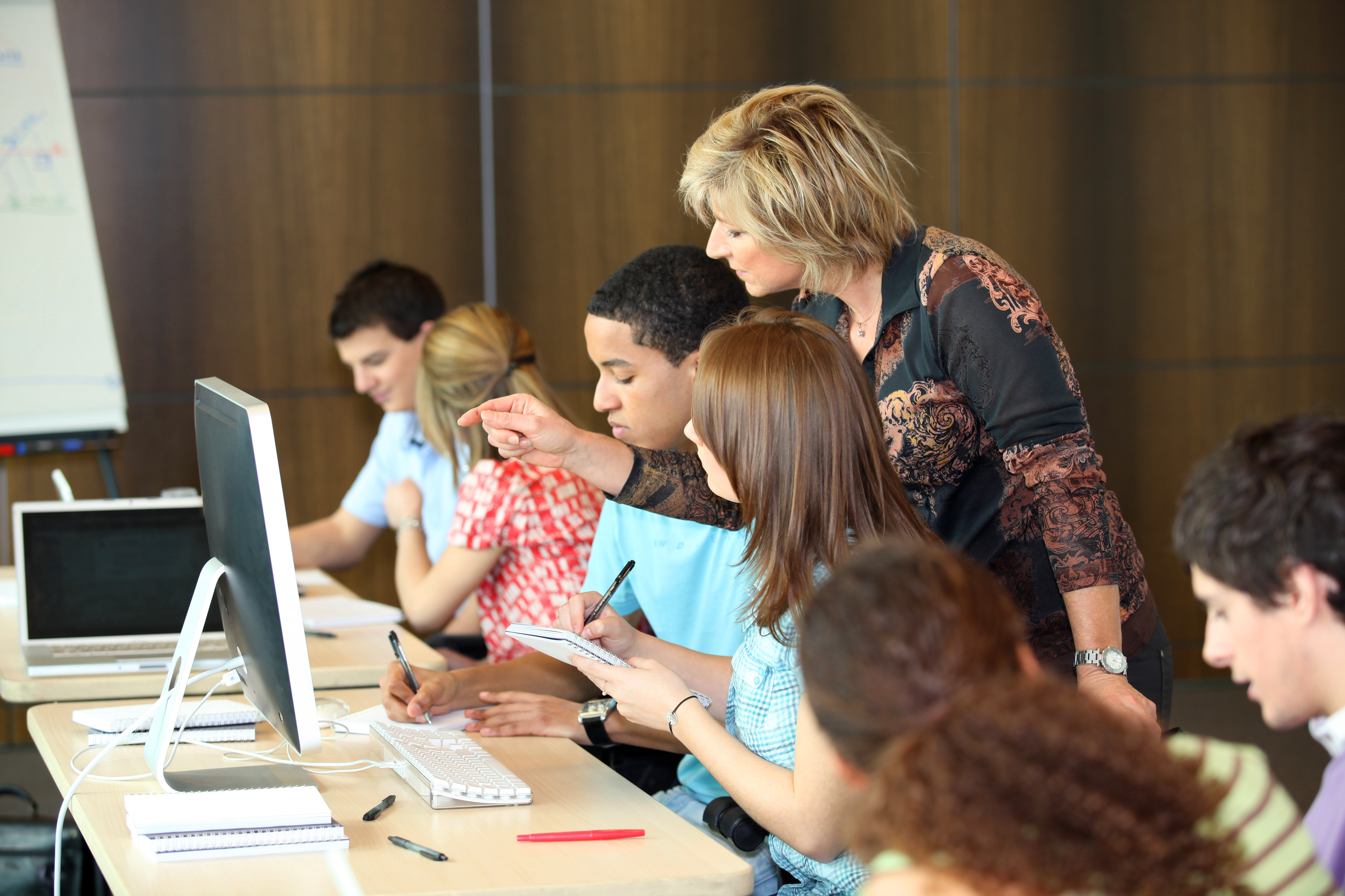 A teacher stands behind several students assisting them with a solution to a problem.