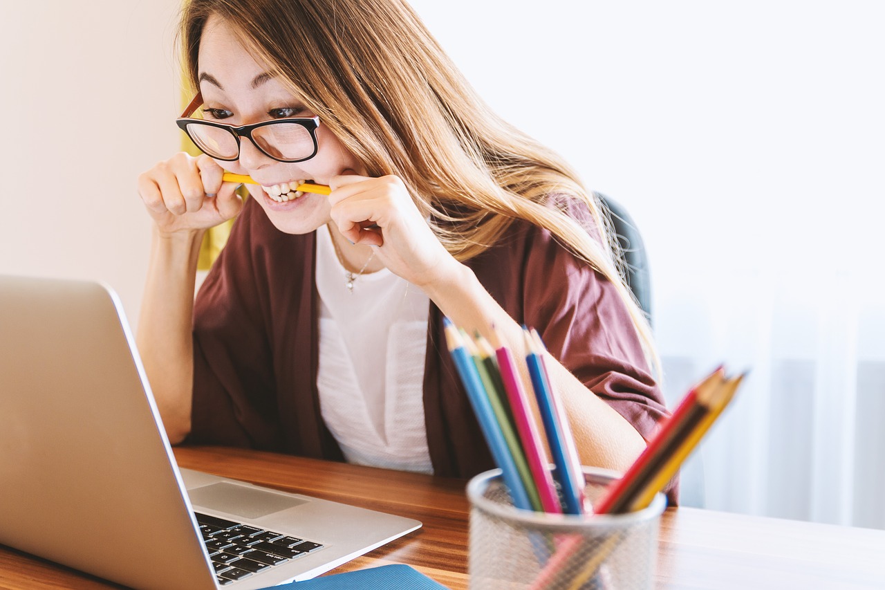 a woman bites a pencil while looking at a laptop