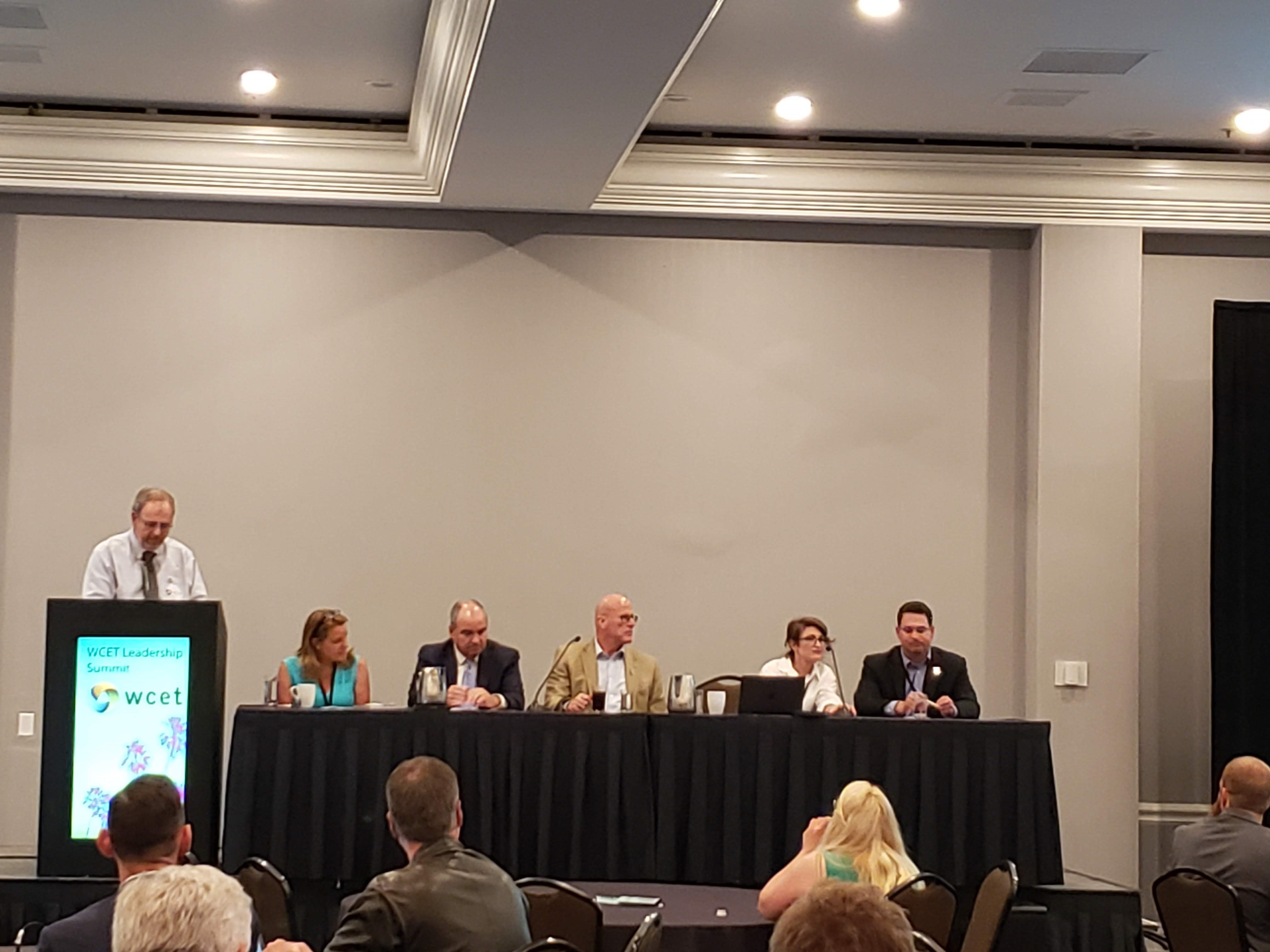 several presenters sit at a long table in front of an audience of attendees. One presenter stands at a podium.