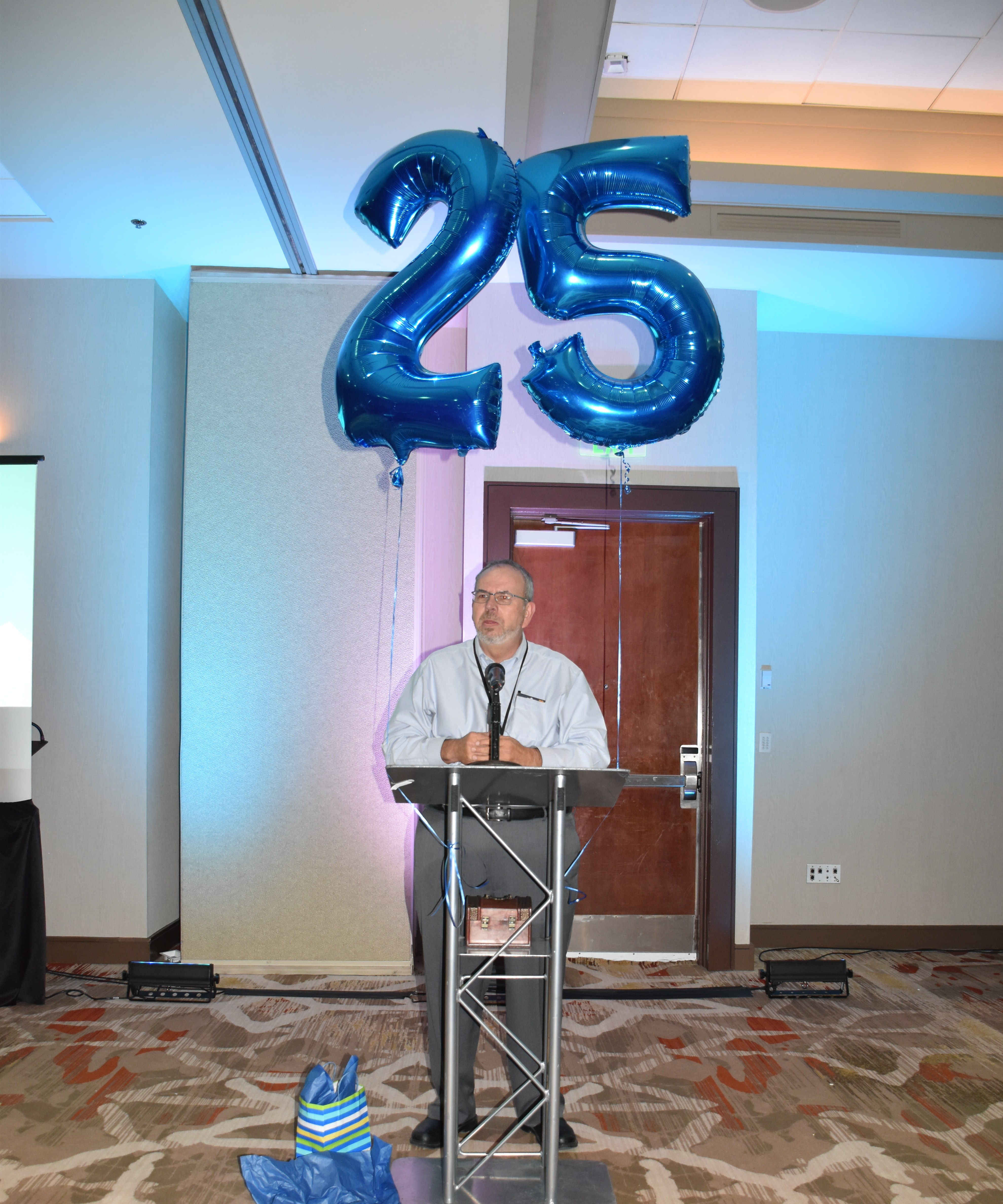 Russ Poulin addressing room with large balloons that say 25