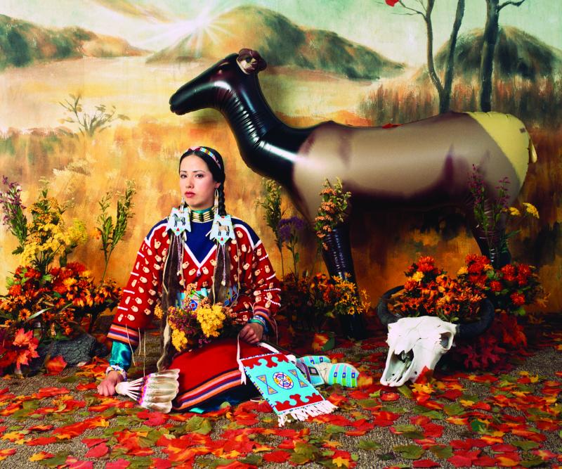 Woman in Native outfit sitting in front of a plains background with a cow skull next to her and an inflatable elk behind her. 