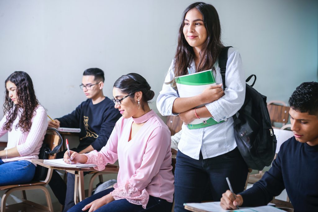 Several college students in class, one standing up and holding notebooks.