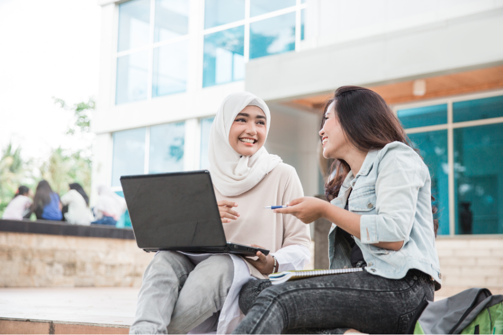 two women sit in front of a building discussing something that is on a laptop in front of them.